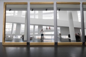 A photo of three very large gold-rimmed mirrors leaning against a wall, with a white building reflected in the background. The photographer’s reflection is centered in the middle.