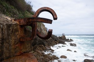 A photo of two parts of a large iron sculpture by artist Eduardo Chillida. The sculptures sit on the coastline with the sea to the right. One piece dominates in the foreground, with another just visible in the background.