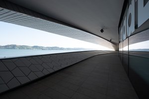 A photo taken from the exterior walkway of the new Maat gallery. On the right is the glass exterior wall of the gallery with the letters ‘maat’ visible in large white text. On the left the Tagus river and shore of Porto Brandão can be seen through a long cut away in the walkway.