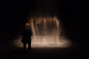 A fine mist of water drops in a dark room, lit from the side. A woman stands in front of the artwork.