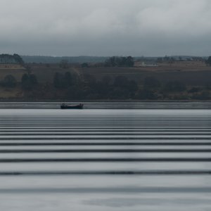Looking across a wide lake on a grey day. In the distance is a small industrial boat. The water has regular horizontal dark bands - like a barcode - caused by ripples from a passing boat.